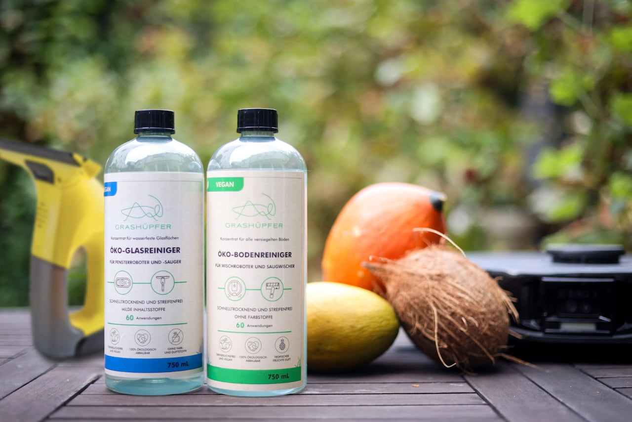 Öko-Glasreiniger und Öko-Bodenreiniger von Grashüpfer stehen auf einem Holztisch, dahinter ist ein Saugroboter mit Wischfunktion und ein Fenster-Sauger und Früchte symbolisieren einen fruchtigen und schönen Duft.