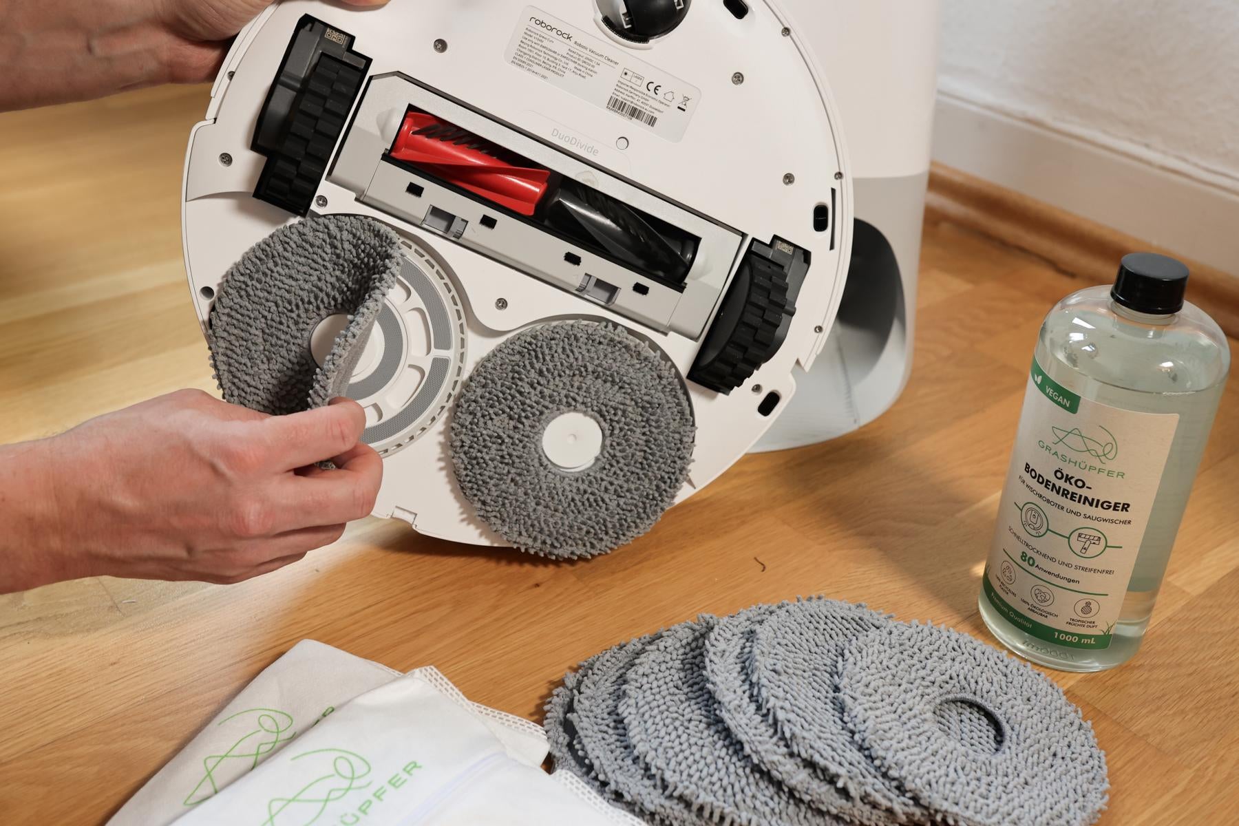 Die Wischpads des Grashüpfer Roborock Sorglospakets werden an einem Roborock Qrevo Modell befestigt und man sieht die perfekte Passform, optimale Kompatibilität und Haftung, daneben sind mehr Wischpads, der Öko-Bodenreiniger für Saugroboter und Staubbeutel als Teil des Sorglospakets zu sehen.