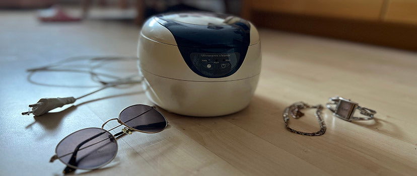 Ein Ultraschallreinigungsgerät liegt auf dem Boden neben einer Brille, einer Uhr und einer Kette.