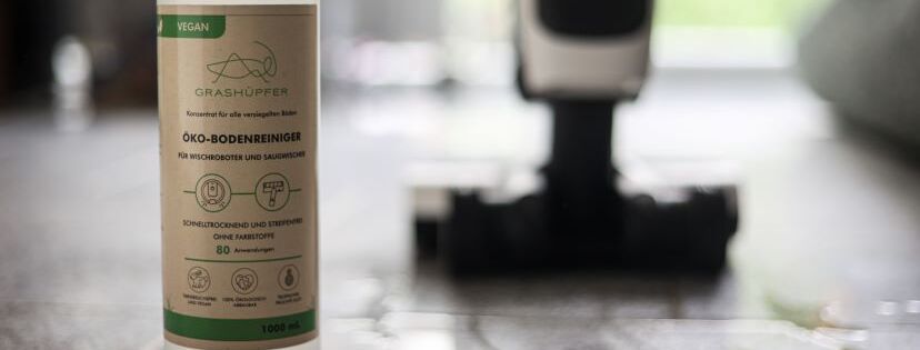 Der Grashüpfer Bodenreiniger für Wischroboter und Saugwischer steht in einer Flasche, als beste, effektive Reinigungsmittel Alternative für Vowerk Geräte neben einem Vorwerk Saugwischer.