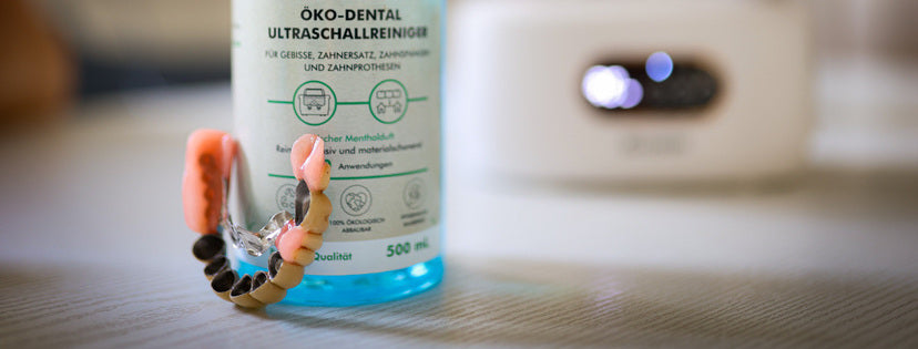 Der Grashüpfer Öko-Dental-Ultraschallreiniger für Zahnprothesen und Co. steht neben einer Zahnprothese und im Hintergrund steht ein Ultraschallreinigungsgerät um Zahnstein von einer Zahnprothese entfernen zu können.