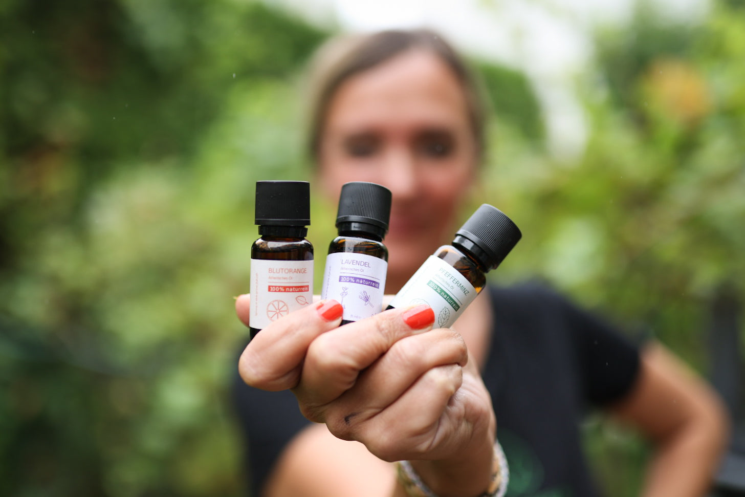 Die Grashüpfer 100% naturreinen ätherischen Öle, Blutorange-, Lavendel- und PFefferminzöl werden von einer Frau in einer Hand in dr Natur hochgehalten.