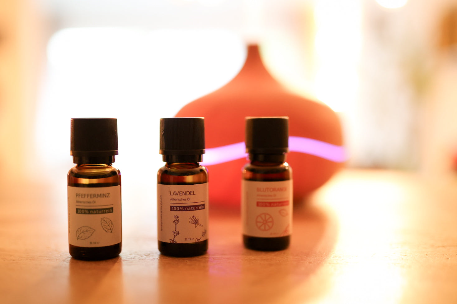 Das 100% naturreine, ätherische Aroma Wohlfühlset von Grashüpfer, bestehend aus Pfefferminzöl, Lavendelöl und Blutorangenöl steht vor einem Aroma Diffuser aufgereiht in den drei Fläschchen.