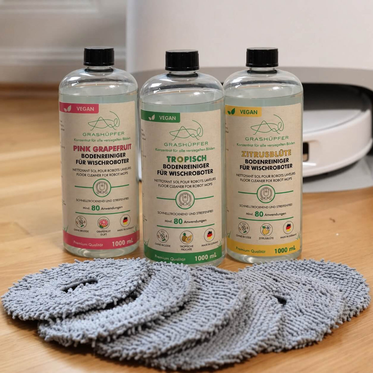 GRASHÜPFER Bodenreiniger Pink Grapefruit, Tropisch und Zitrusblüte für Wischroboter mit mehreren grauen Wischpads vor einem Roborock Saug-Wischroboter.