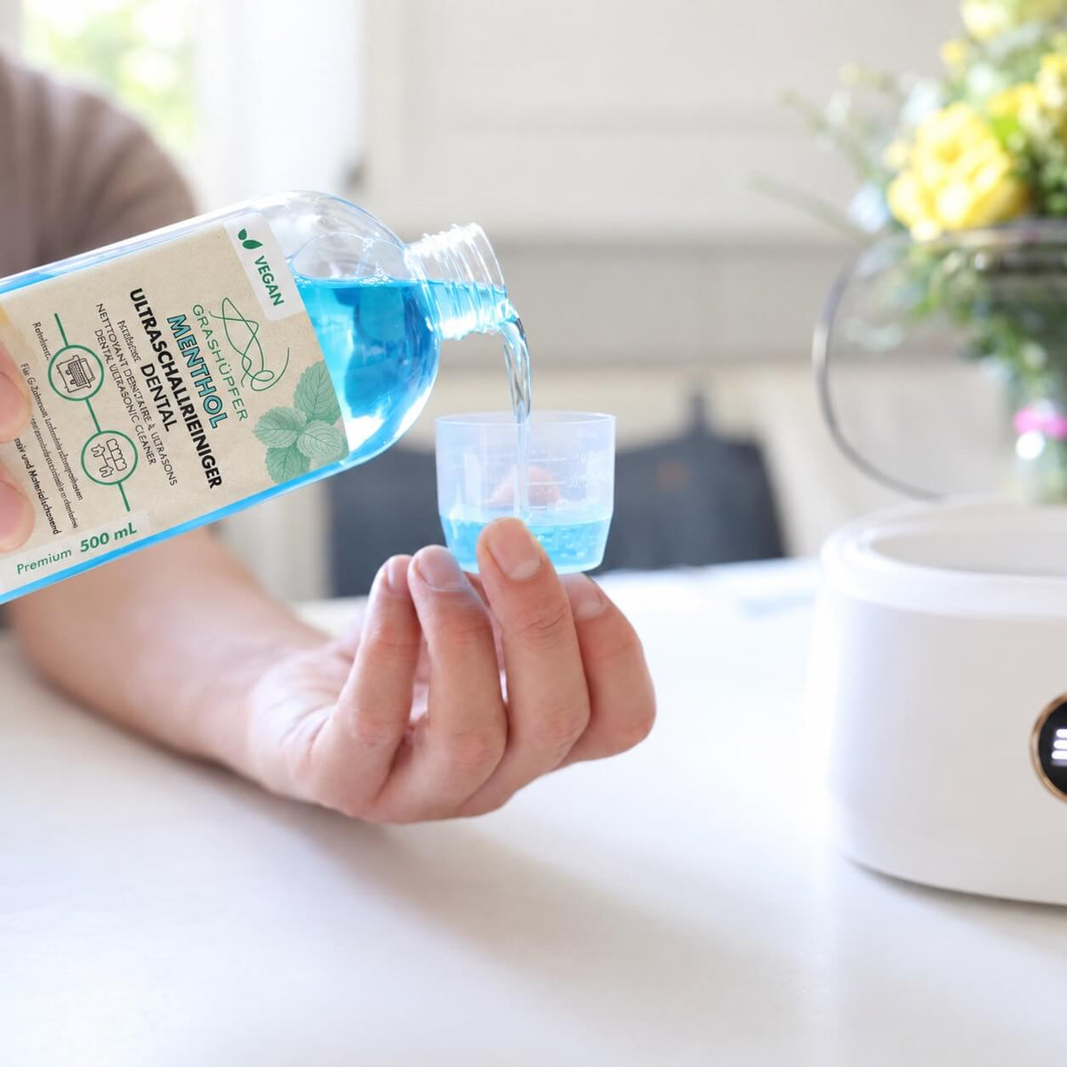 GRASHÜPFER Dental Ultraschallreiniger Menthol wird zur Dosierung in einen Messbecher neben einem Ultraschallreinigungsgerät gegossen.