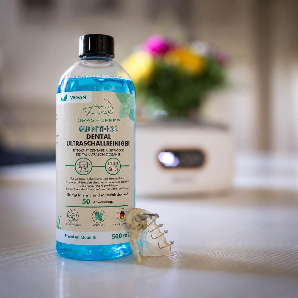 GRASHÜPFER Dental Ultraschallreiniger Menthol 500 ml neben herausnehmbarem Zahnersatz und einem Ultraschallreinigungsgerät.