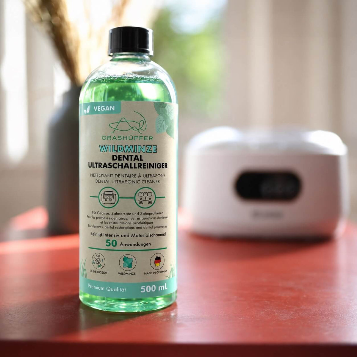GRASHÜPFER Dental Ultraschallreiniger Wildminze 500 ml vor einem Ultraschallreinigungsgerät zur Reinigung von Zahnersatz und Zahnprothesen.