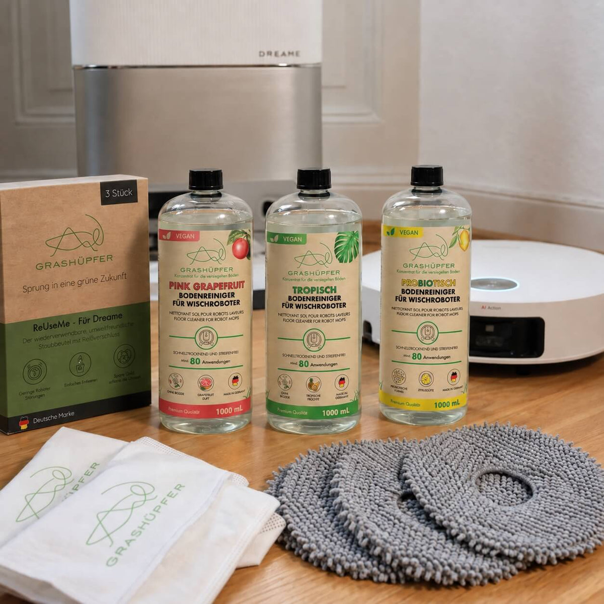 Grashüpfer Dreame Mix & Match Set mit Bodenreiniger, probiotischem Bodenreiniger, ReUseMe-Staubbeuteln und grauen Wischpads vor einem Dreame Saug-Wischroboter mit Station.