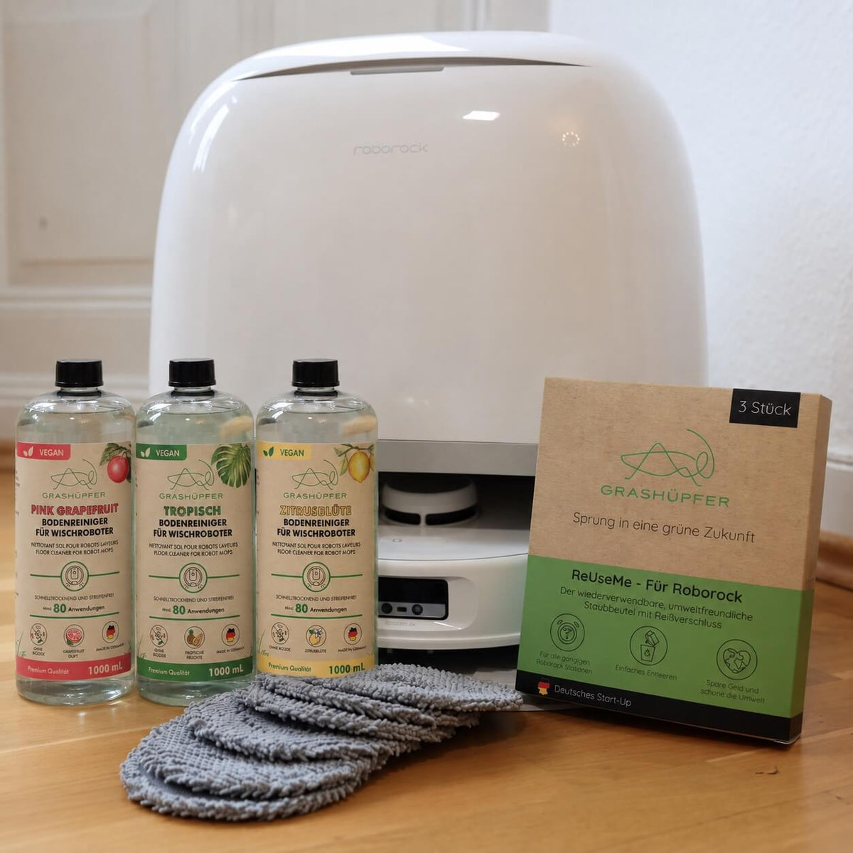 GRASHÜPFER Roborock Zubehör mit Bodenreinigern Pink Grapefruit, Tropisch und Zitrusblüte, ReUseMe Staubbeuteln und grauen Wischpads vor der Servicestation.