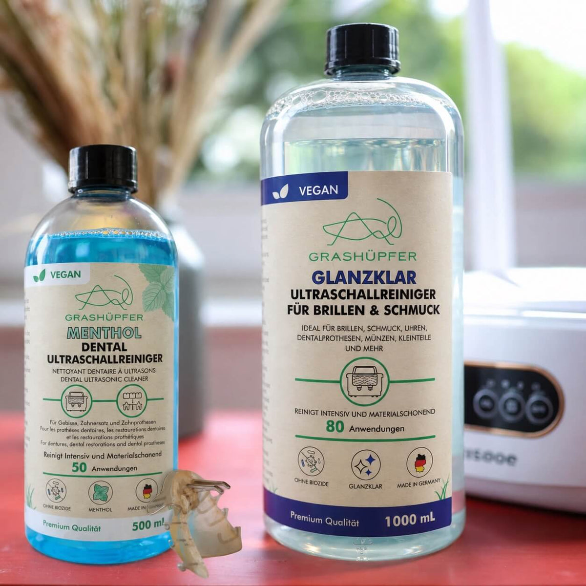 GRASHÜPFER Ultraschallreiniger Duo mit Dental Menthol und Glanzklar, Zahnersatz und Ultraschallgerät im Hintergrund.