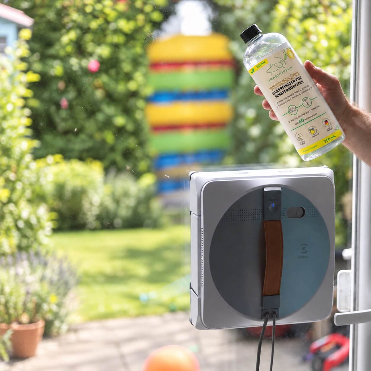 GRASHÜPFER Zitrusblüte Glasreiniger für Fensterroboter wird neben einem Fensterreinigungsroboter an einer Glasscheibe gezeigt.