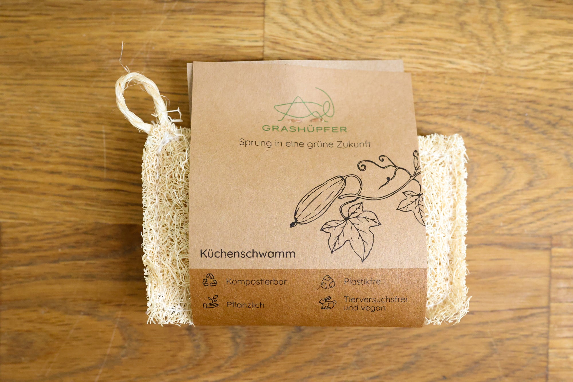 Der Luffa Schwamm von Grashüpfer liegt in der Verpackung auf einem Holztisch und auf der Verpackung stehen positive Eigenschaften wie kompostierbar, plastikfrei, pflanzlich, tierversuchsfrei und vegan.