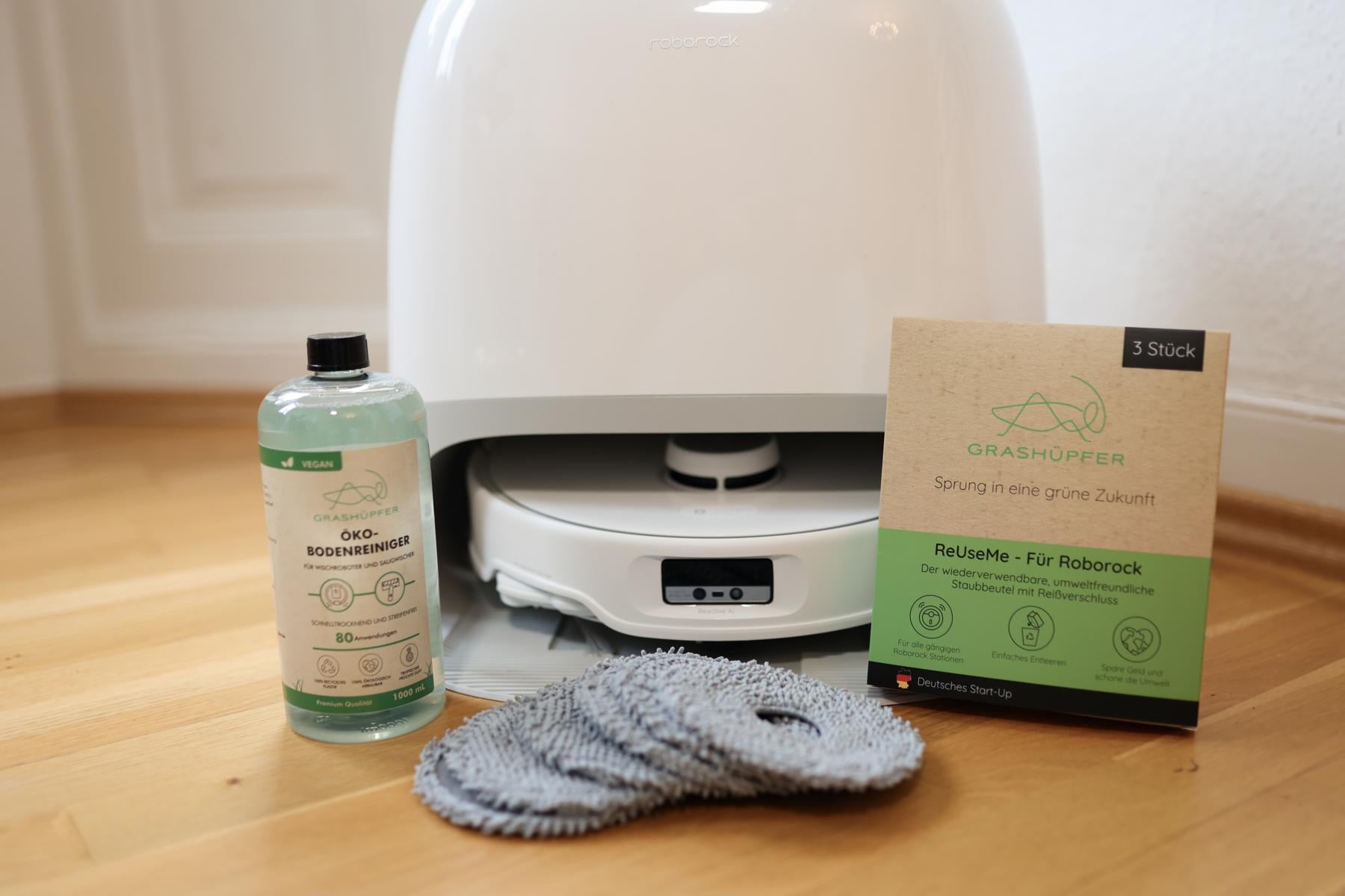 Das Roborock Sorglospaket von Grashüpfer liegt in einzelteilen da, darunter eine Flasche des Öko-Bodenreinigers, 6 Wischpads und eine Verpackung der ReUseMe, wiederverwendbaren Staubbeutel für Roborock Service-Stationen.