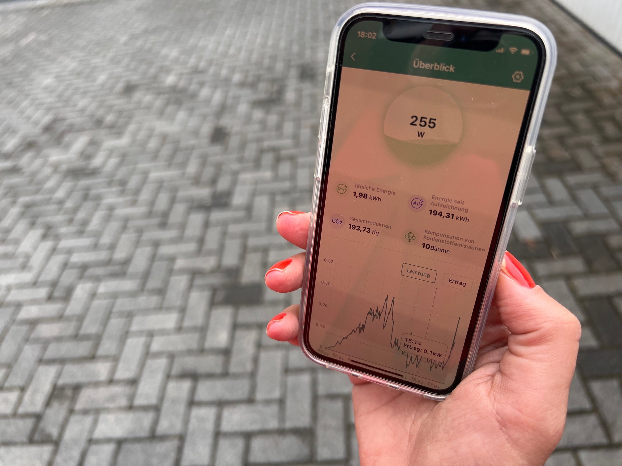 Eine Frauenhand mit roten Fingernägeln hält ein Smartphone in der Hand, welches die aktuelle Solarproduktion zeigt, welche durch die Reinigung des Solarmoduls mit dem Öko-Solarreiniger von Grashüpfer deutlich verbessert wurde.